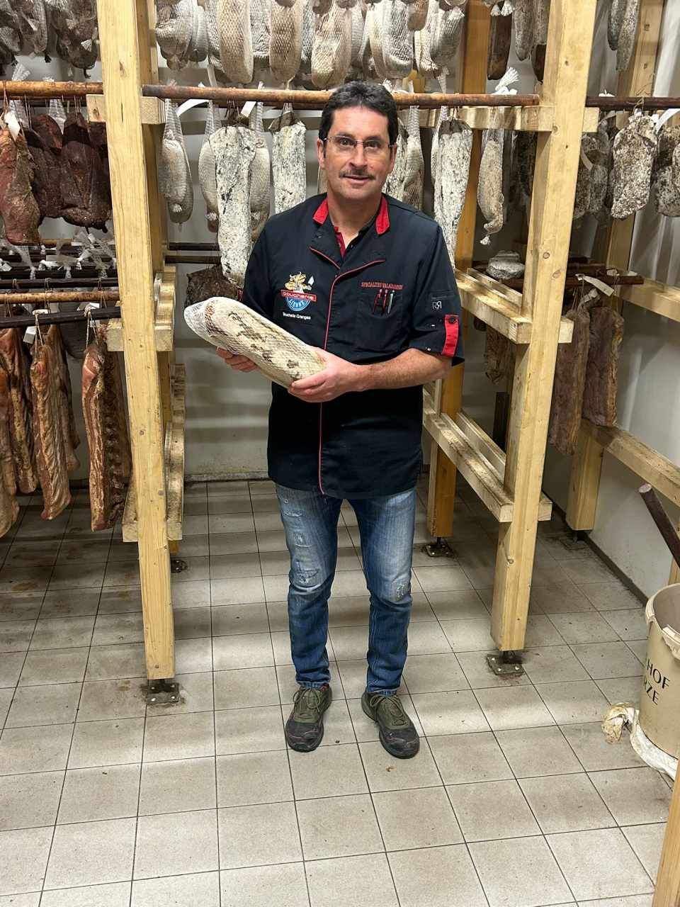 Un homme tient de la charcuterie dans une réserve où sont suspendues des rangées de charcuteries.