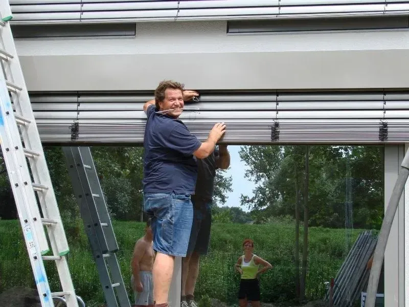 Ein Mann montiert einen Rollladen an einem Gebäude. Zwei weitere Personen stehen in der Nähe.