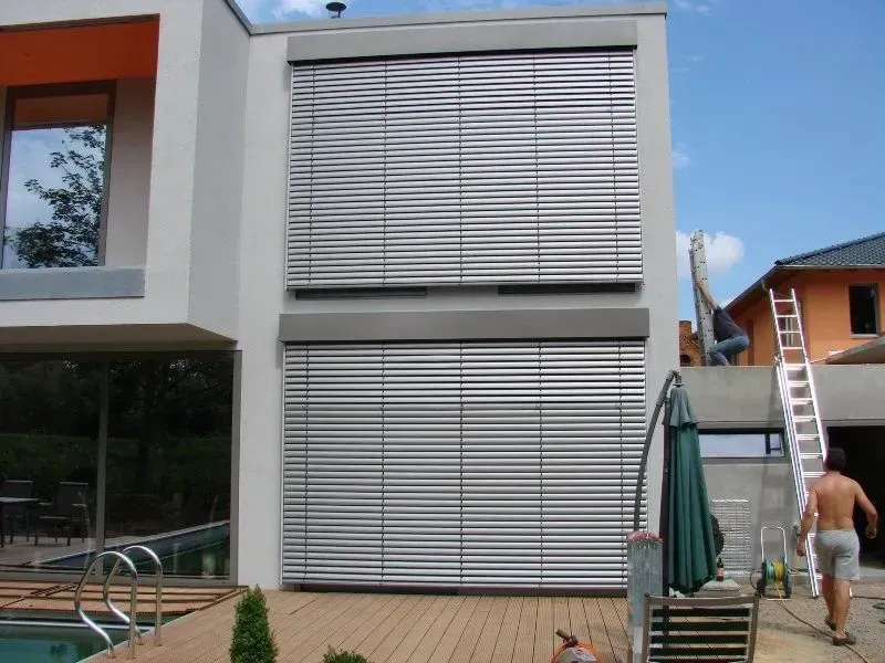 Außenansicht eines modernen Hauses mit großen Jalousien. Eine Person steht neben einer Leiter. Pool und Terrasse sind zu sehen.