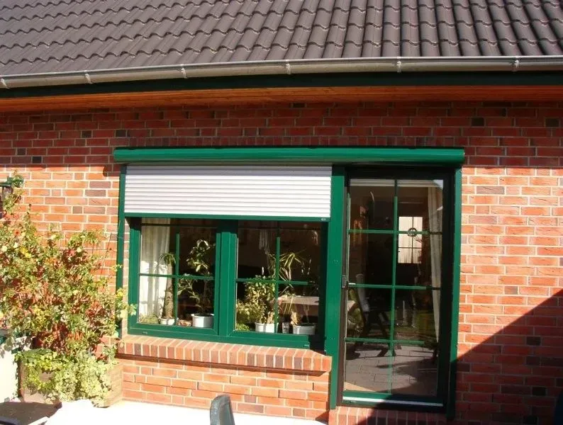 Fenster und Tür mit grünem Rahmen und geschlossenem, hellfarbigem Rollladen an einem roten Backsteingebäude.