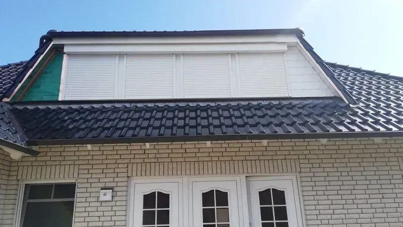 Weiße Fensterläden an einem Haus, über einer Backsteinfassade und einem dunklen Ziegeldach, unter einem klaren blauen Himmel.