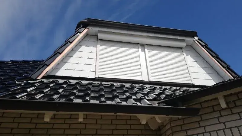 Weiße Dachfensterläden an einem Dachgaubenfenster, schwarzes Ziegeldach, blauer Himmel.