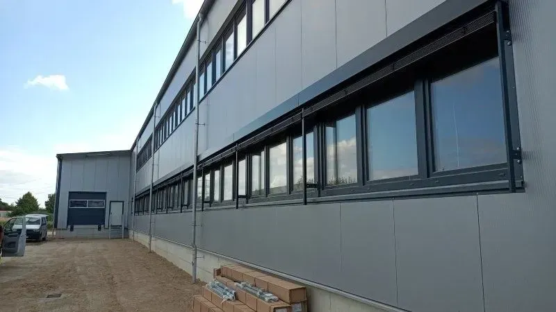 Graues Industriegebäude mit zahlreichen Fenstern und Außenjalousien unter blauem Himmel.