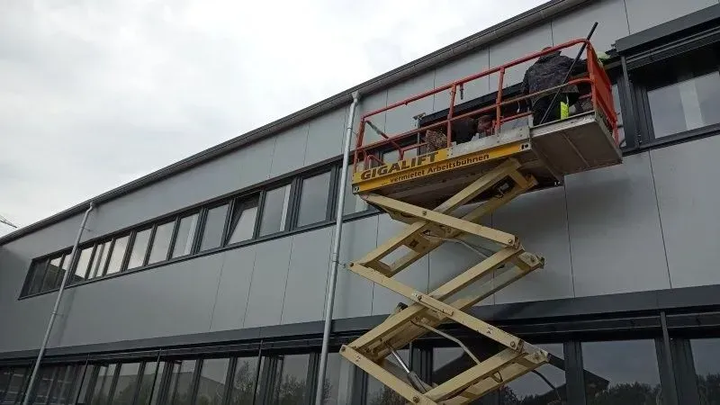 Ein Arbeiter ist auf einer Scherenbühne bei Arbeiten an der Außenfassade eines Gebäudes mit grauer Verkleidung und dunklen Fenstern tätig.