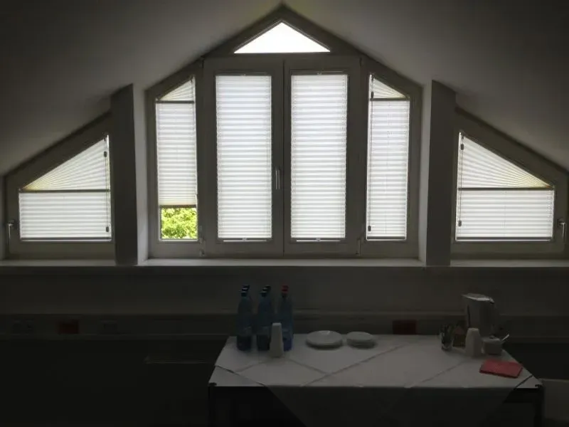 Dreieckiges Fenster mit Jalousien, Flaschen und gedecktem Tisch in einem schwach beleuchteten Raum.