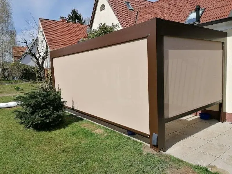 Braune Pergola mit beigen Stoffschirmen in einem Garten, Haus im Hintergrund.