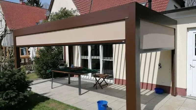 Braune und beige Terrassenpergola mit eingefahrenem Sonnenschutz über einer Terrasse mit Tisch und Stühlen.