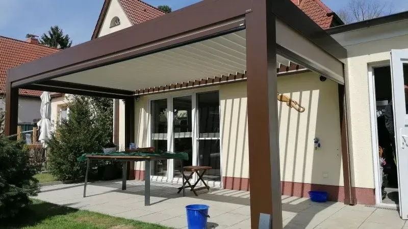 Eine braune Pergola spendet Schatten über einer Terrasse mit Schiebetüren; ein kleiner Tisch und ein blauer Eimer sind zu sehen.