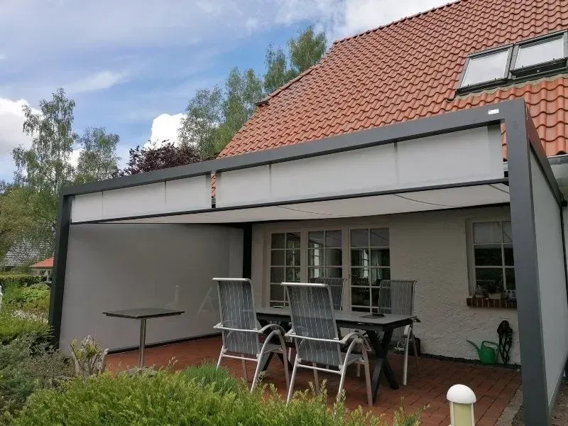 Terrasse mit grauer Pergola, weißen Sonnenschirmen, Tisch und Stühlen, angrenzend an ein Backsteinhaus.