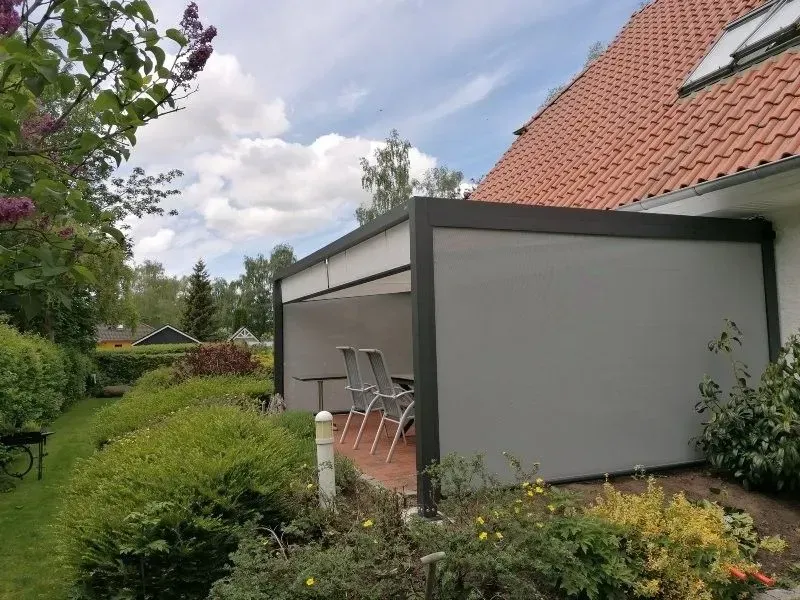 Graue Terrassenüberdachung an einem Haus mit rotem Dach. Weiße Decke, graue Wände, Gartenmöbel und üppige grüne Bepflanzung.