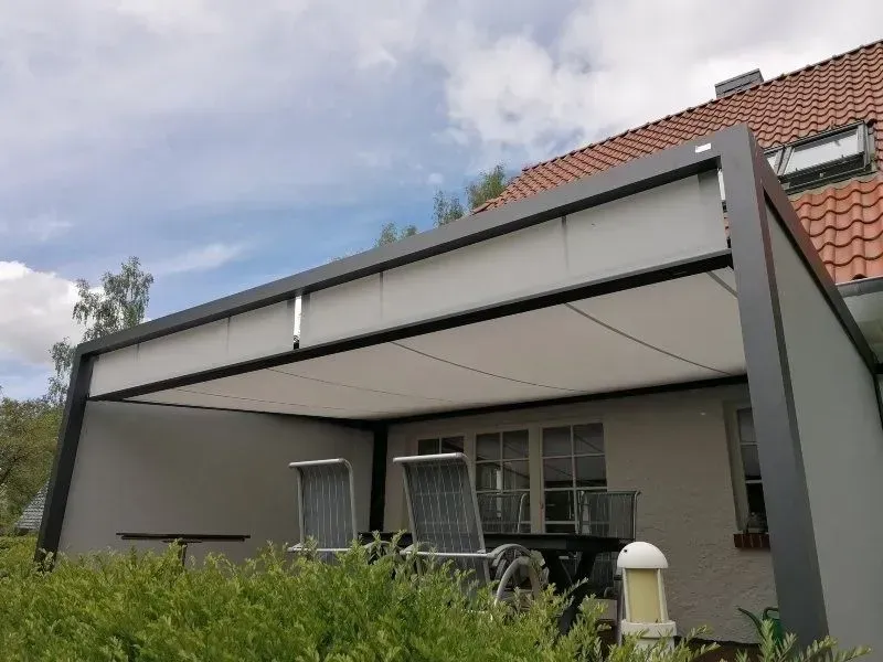 Graue Terrassenmarkise über Terrassenmöbeln vor einem weißen Haus mit orangefarbenem Ziegeldach, bewölkter Himmel.