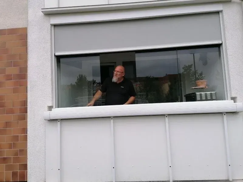 Ein Mann mit Brille lehnt sich aus einem Fenster mit hellgrauer Markise, weißem Balkongeländer und Backsteinmauer.