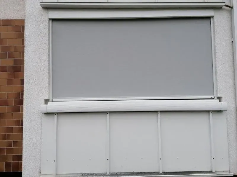 Ein grauer Rollo verdeckt teilweise ein weiß gerahmtes Fenster an einem Gebäude mit Backsteinmauer.