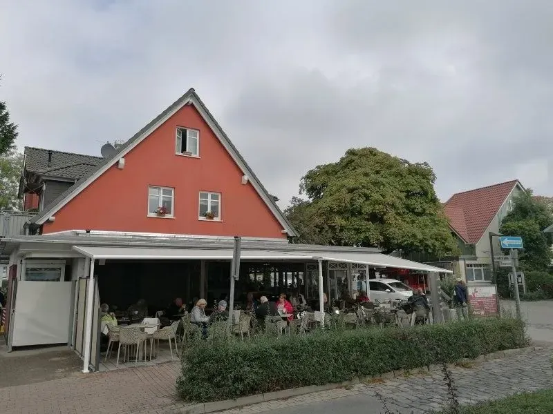 Café mit Außensitzplätzen unter einer weißen Markise, neben einem roten Gebäude. Die Gäste sitzen an Tischen.