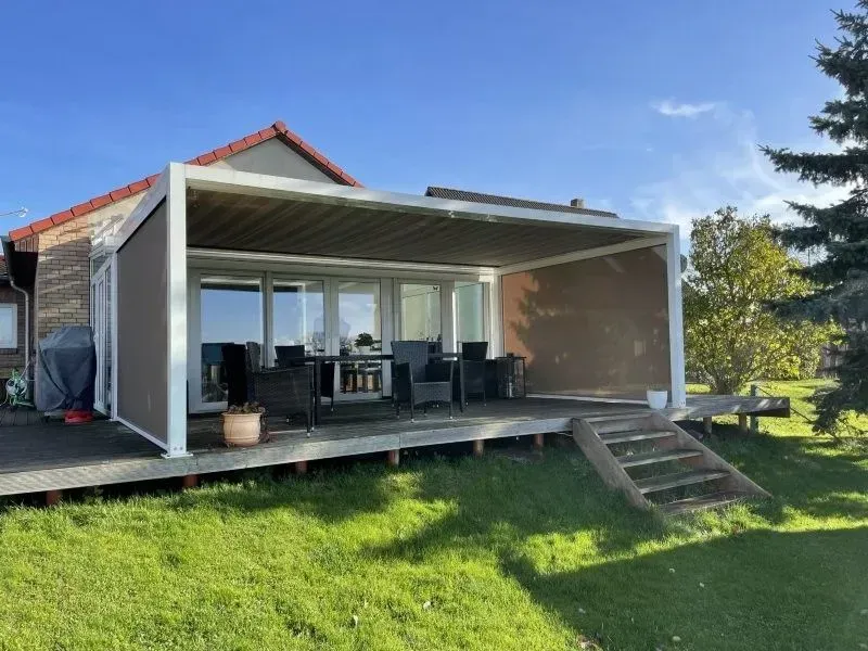 Terrasse mit Esstisch und Stühlen, überdacht von einer Pergola. Braune Wände und Sonnenschutz, grüner Rasen.