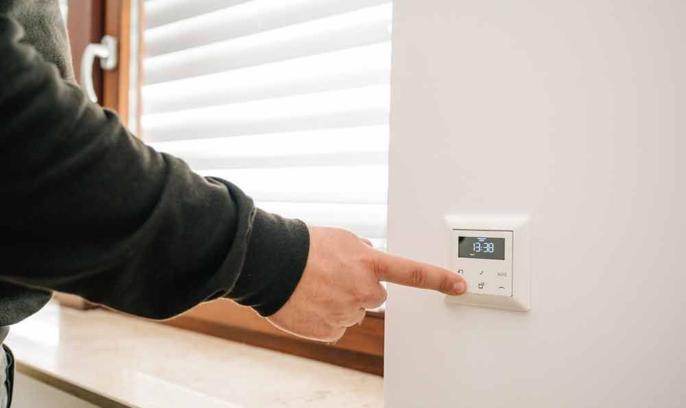 Eine Person drückt mit dem Finger einen Knopf an einem weißen, an der Wand montierten Thermostat. Im Hintergrund ist ein Fenster mit Jalousien zu sehen.