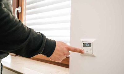 Eine Person drückt mit dem Finger einen Knopf an einem weißen, an der Wand montierten Thermostat. Im Hintergrund ist ein Fenster mit Jalousien zu sehen.