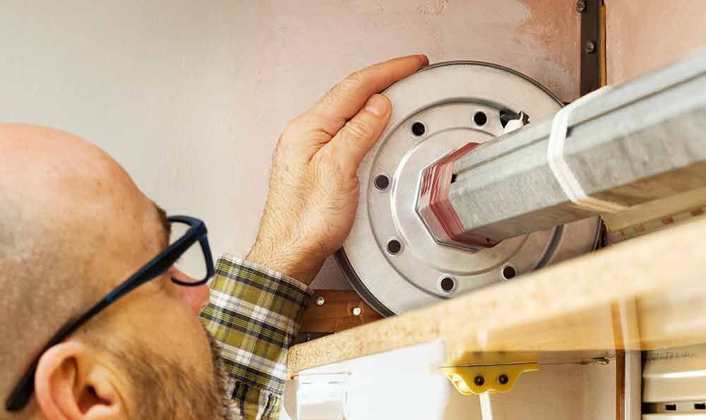 Mann mit Brille arbeitet an einem Rollladenmechanismus, Innenaufnahme.