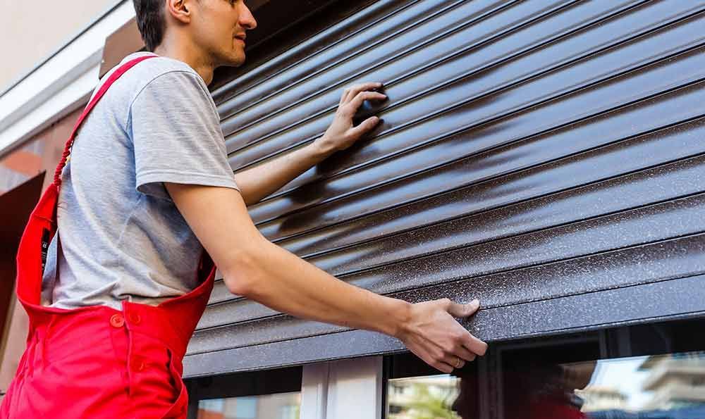 Ein Mann in rotem Overall montiert einen dunklen Metallrollladen an einem Fenster.