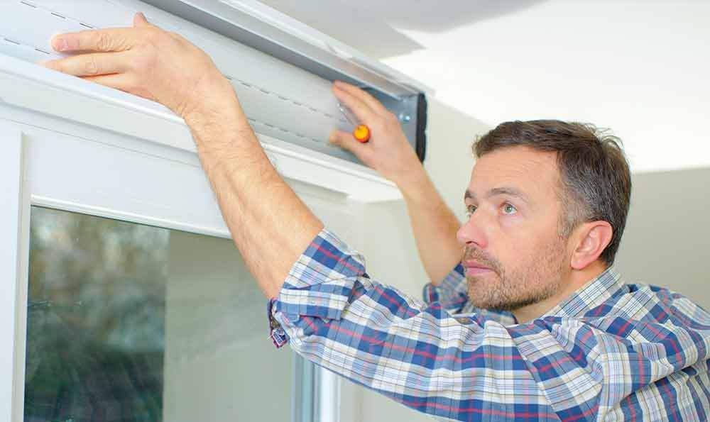 Ein Mann montiert eine Jalousie. Er trägt ein kariertes Hemd und benutzt einen Schraubenzieher. Innenaufnahme.