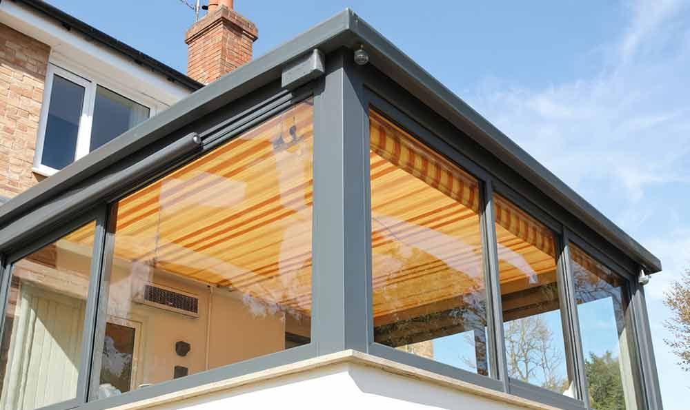 Grau umrahmter Wintergarten mit Klarglasscheiben, an ein Backsteinhaus angebaut, teilweise von beigen Jalousien beschattet, vor blauem Himmel.