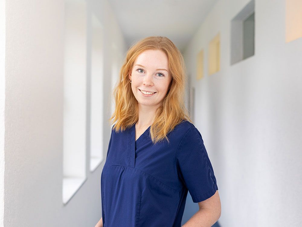 Eine Frau in einem blauen Kittel steht in einem Flur und lächelt.