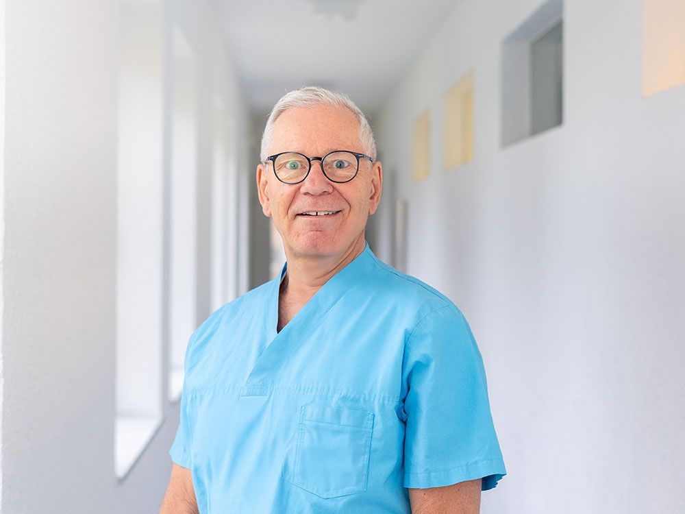 Ein Mann in blauem Kittel und Brille steht in einem Flur.