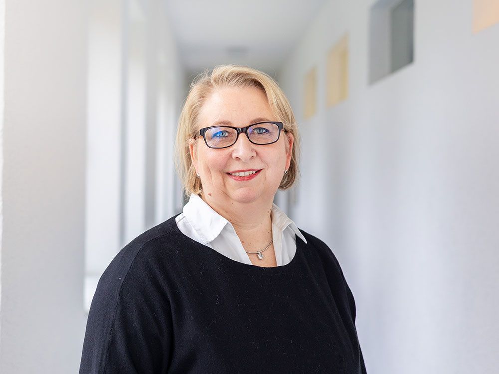 Eine Frau mit Brille und schwarzem Pullover steht in einem Flur.