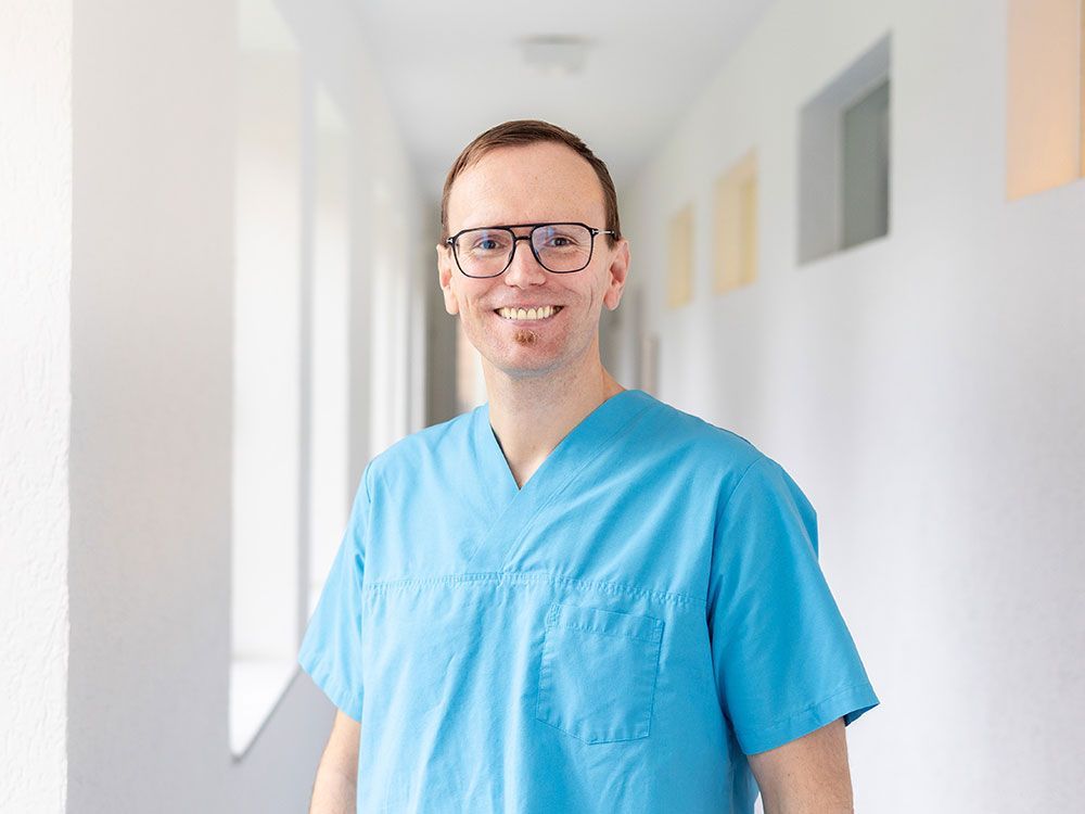 Ein Mann in blauem Kittel und Brille steht in einem Flur.