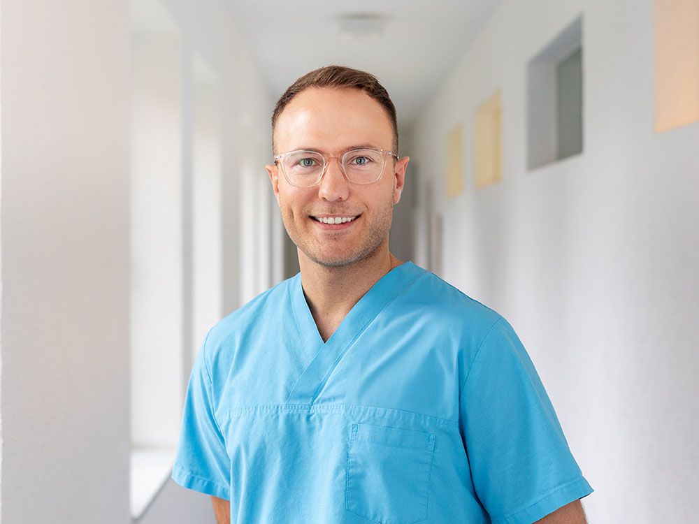 Ein Mann in einem blauen OP-Kittel und mit Brille steht in einem Flur.
