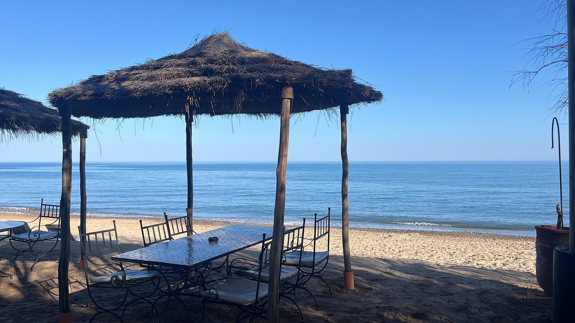 Mesa y sillas junto a la playa bajo estructuras de sombra con techo de paja, con vista al océano bajo un cielo azul claro.