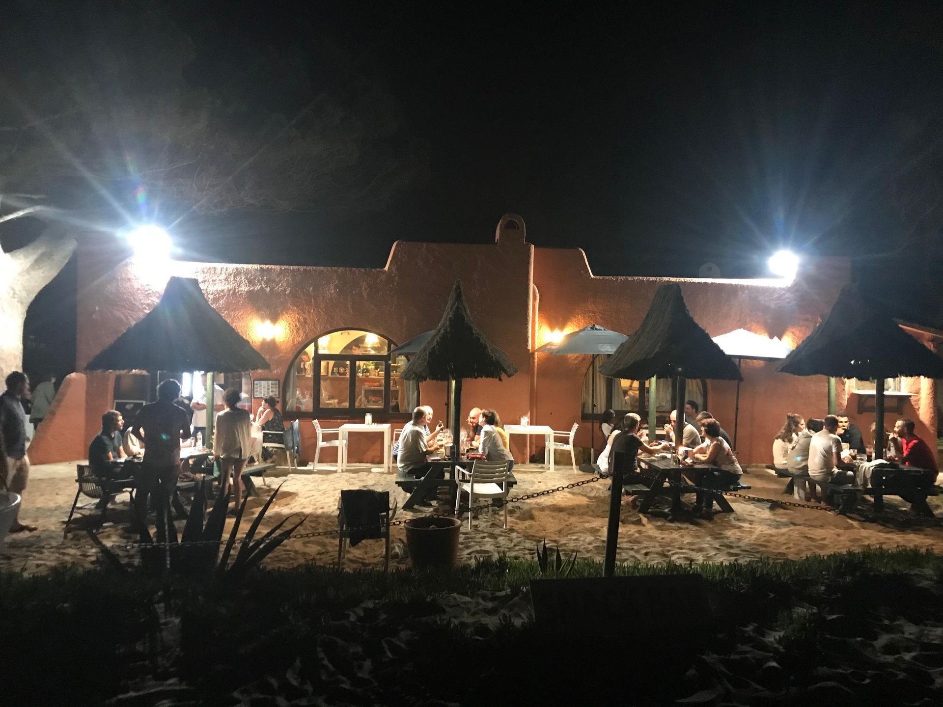 Restaurante al aire libre de noche, con clientes sentados en mesas bajo sombrillas. El edificio es de color terracota.