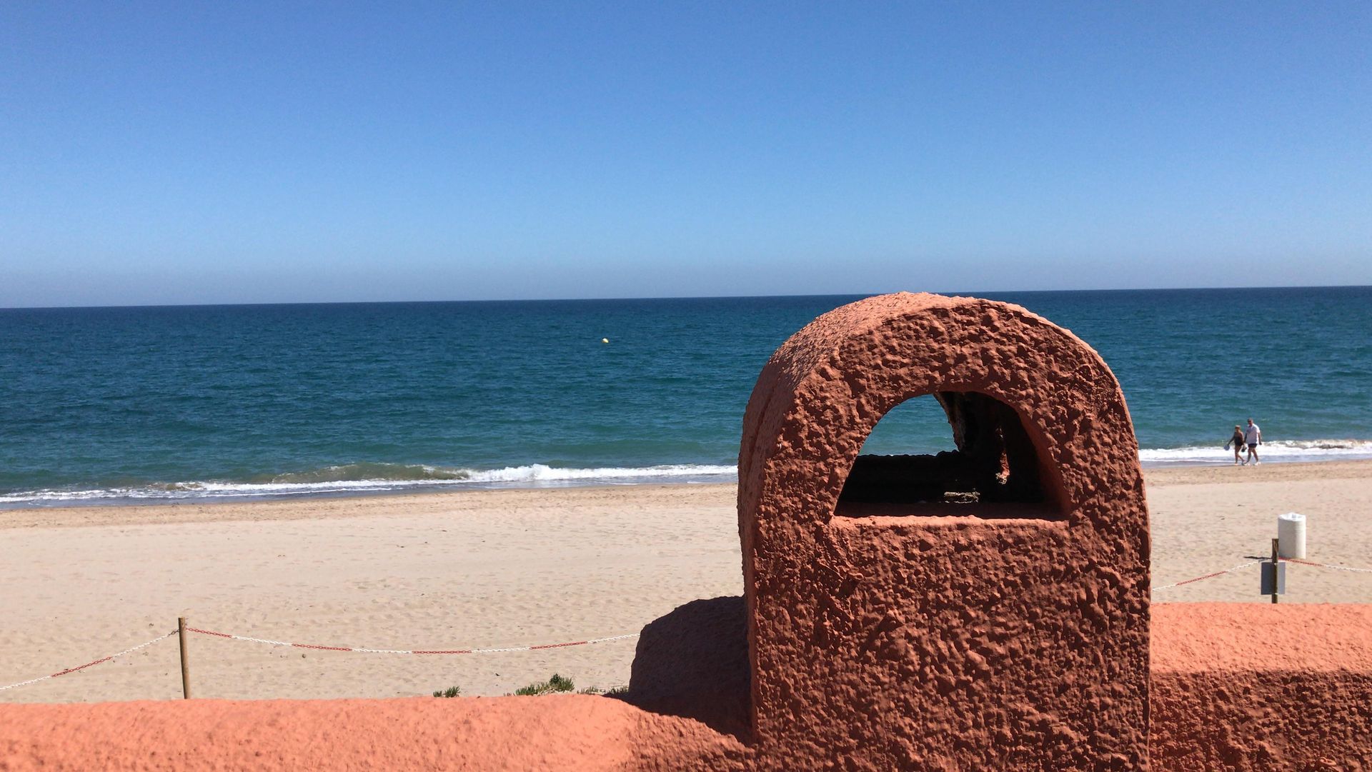 Escena de playa con agua turquesa, orilla de arena y una estructura de color terracota en primer plano.