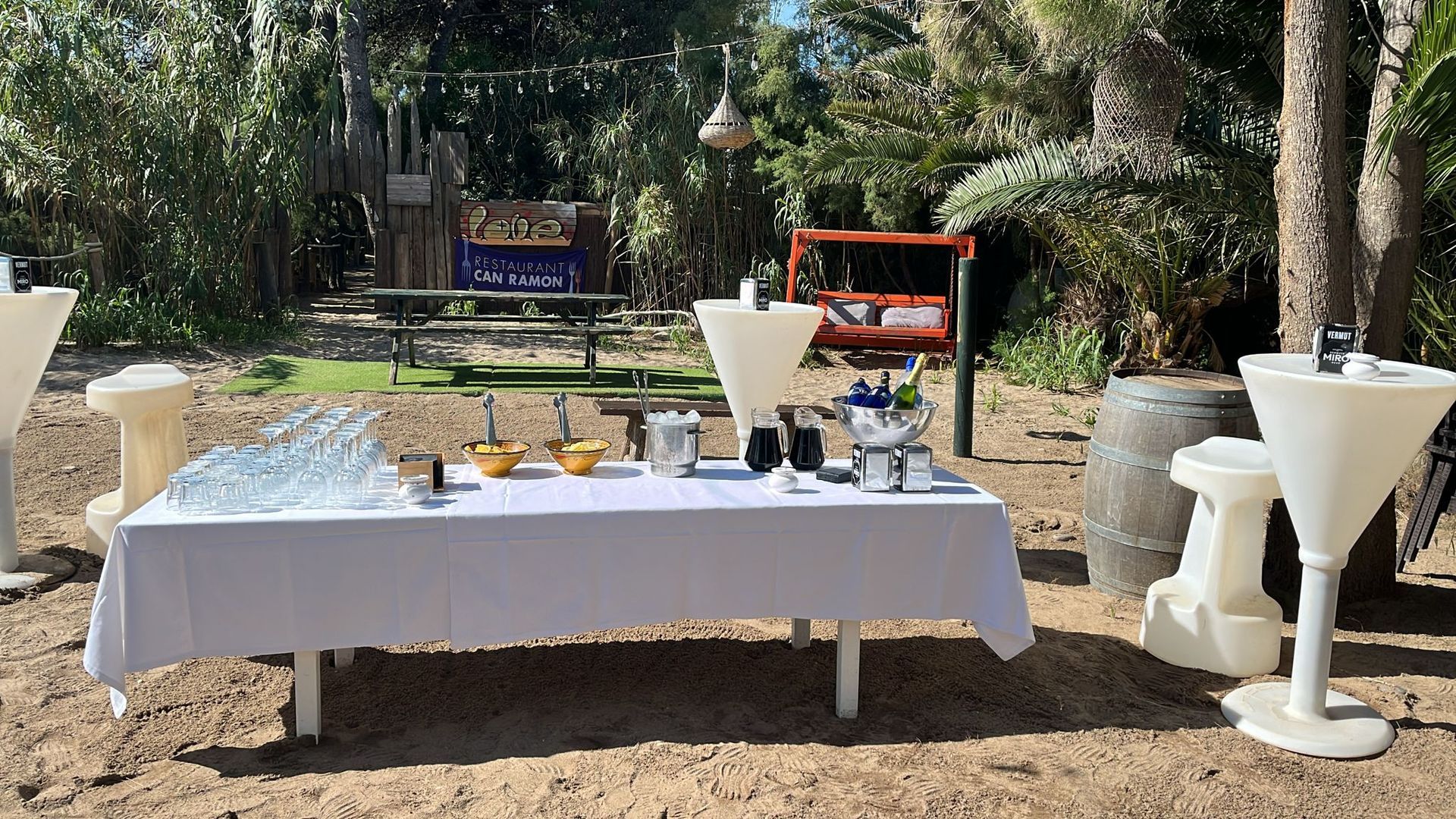 Montaje de catering al aire libre: mesa blanca con comida y bebidas, mesas altas cerca, árboles de fondo.