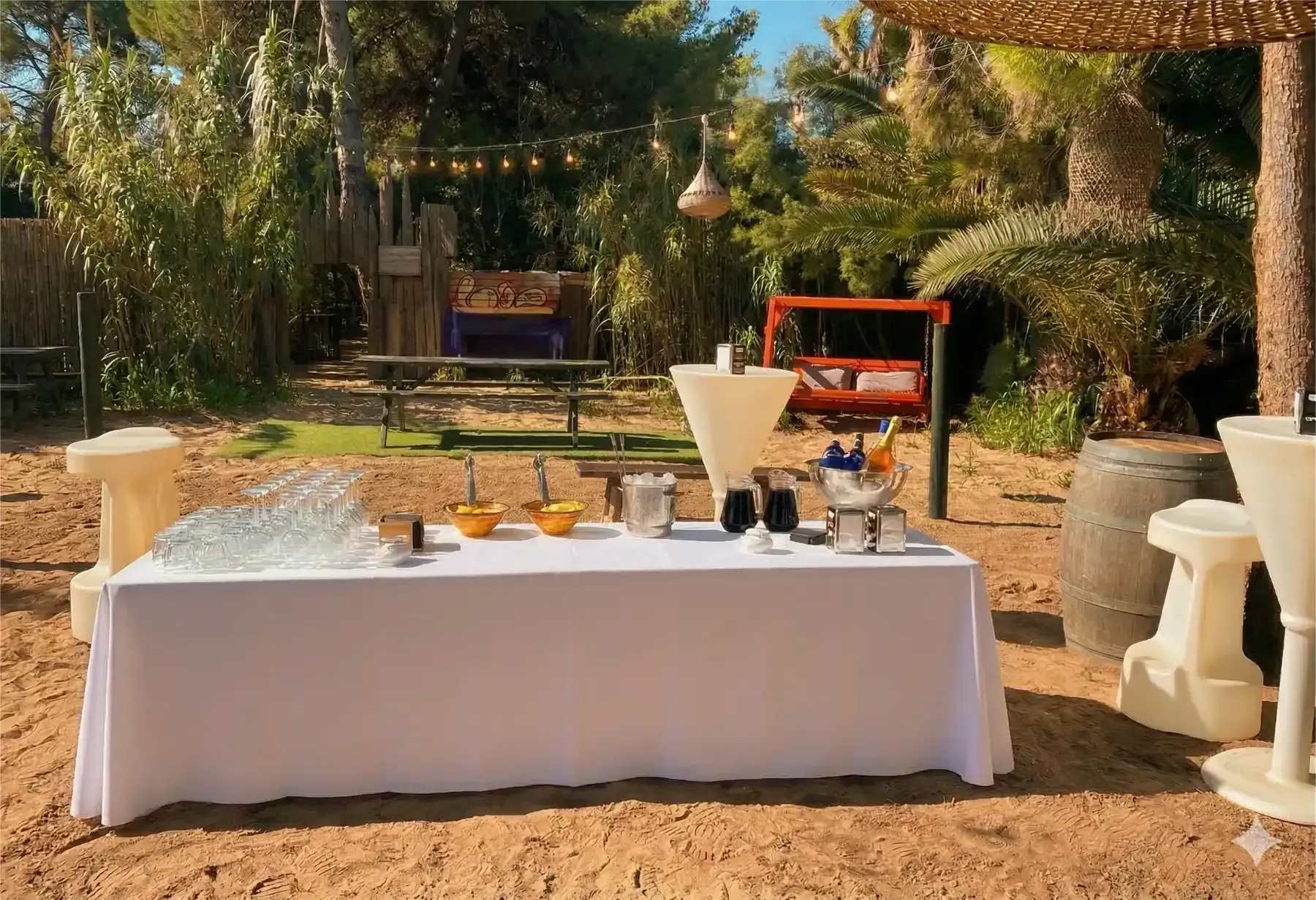 Montaje de catering al aire libre: mesa blanca con comida y bebidas, mesas altas cerca, árboles de fondo.