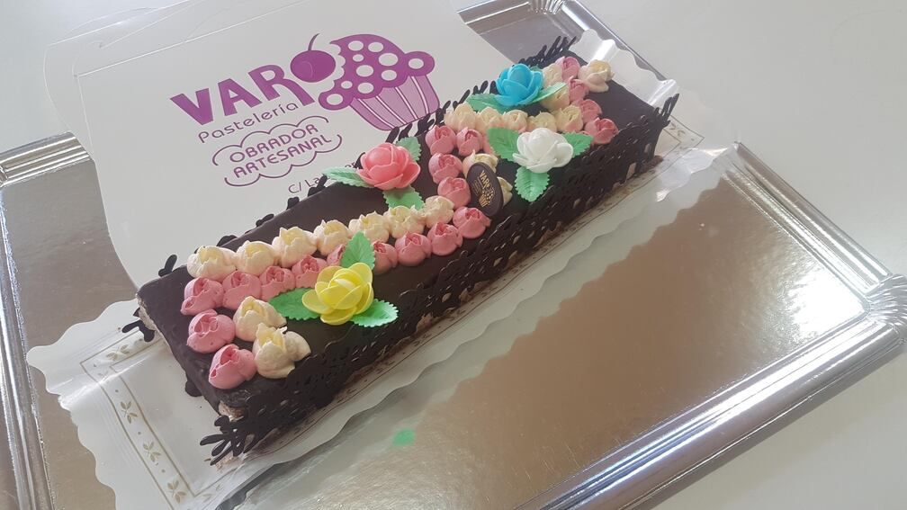 Pastel de chocolate con flores de glaseado de colores sobre una bandeja de plata con el logotipo de una panadería.
