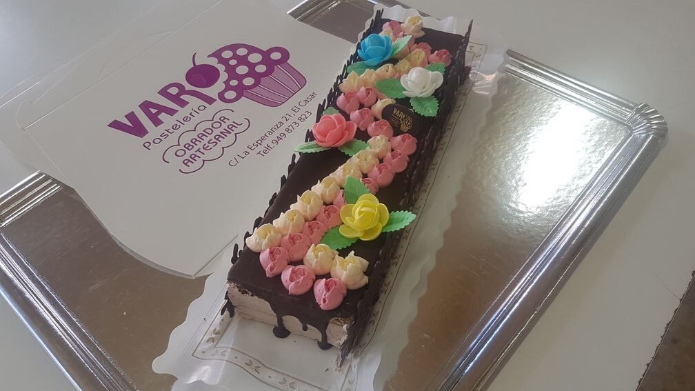 Pastel de chocolate decorado con flores de glaseado de colores en una bandeja de plata. Al fondo, una bolsa de panadería.