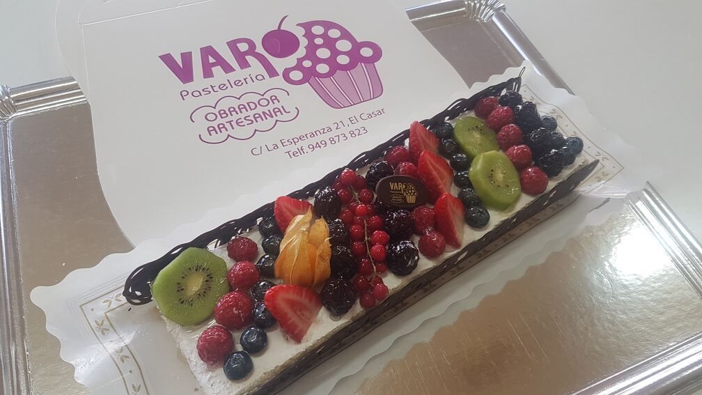 Tarta de frutas con kiwi, frutos rojos y chocolate en una bandeja de plata, junto al logotipo de una panadería.