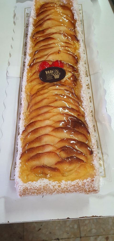 Tarta rectangular de manzana glaseada, coronada con un emblema decorativo y cerezas, sobre papel blanco.