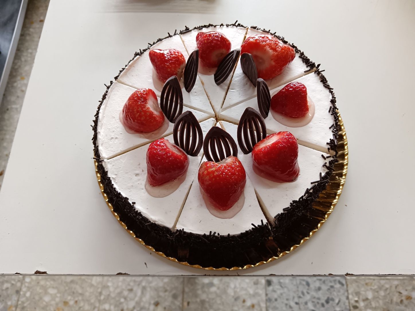 Pastel con glaseado blanco, fresas y decoraciones de chocolate sobre una base dorada.