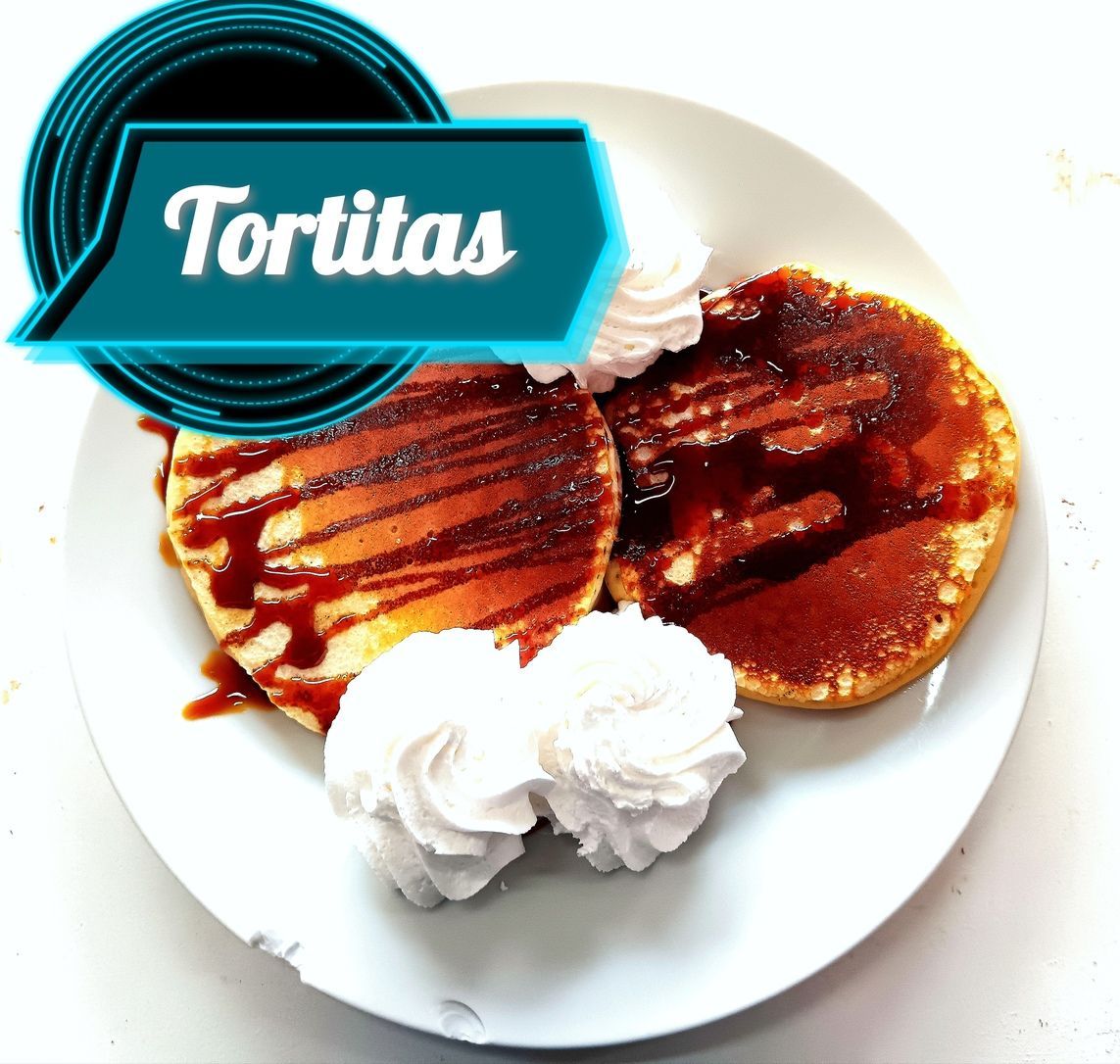 Dos tortitas doradas cubiertas con almíbar y crema batida en un plato blanco.