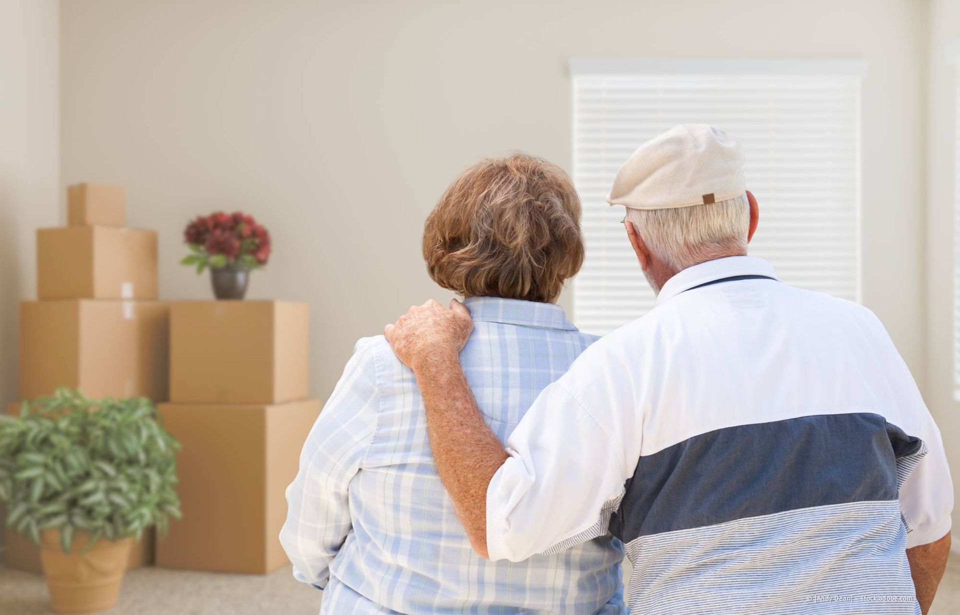 Ein Paar besichtigt ein neues Haus, Umzugskartons und eine Pflanze stehen daneben. Der Mann hat seinen Arm um die Schulter der Frau gelegt.