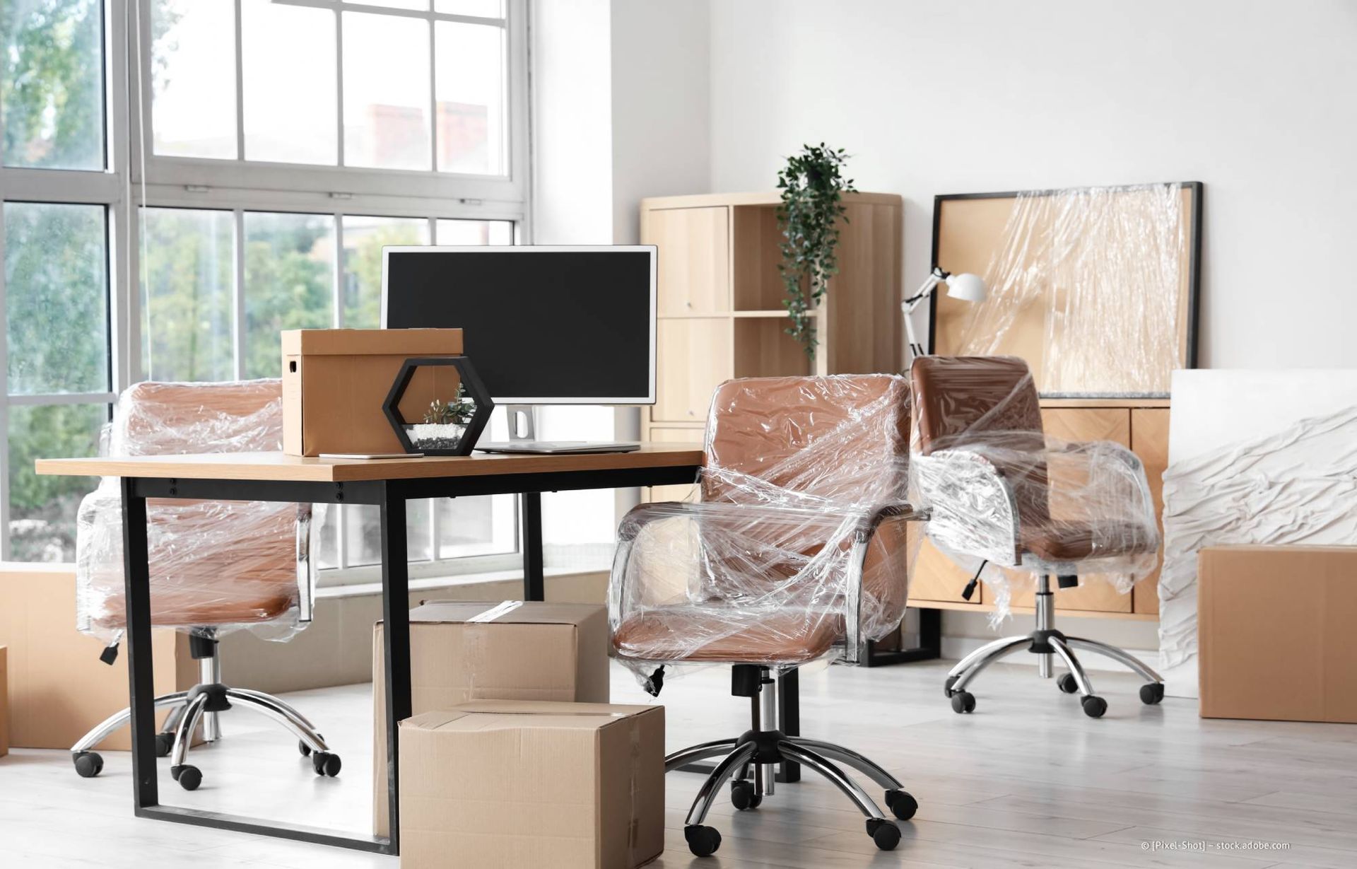 Büroräume werden verlegt. Stühle und andere Gegenstände sind in Plastikfolie verpackt, Kisten und Möbel befinden sich im Raum.