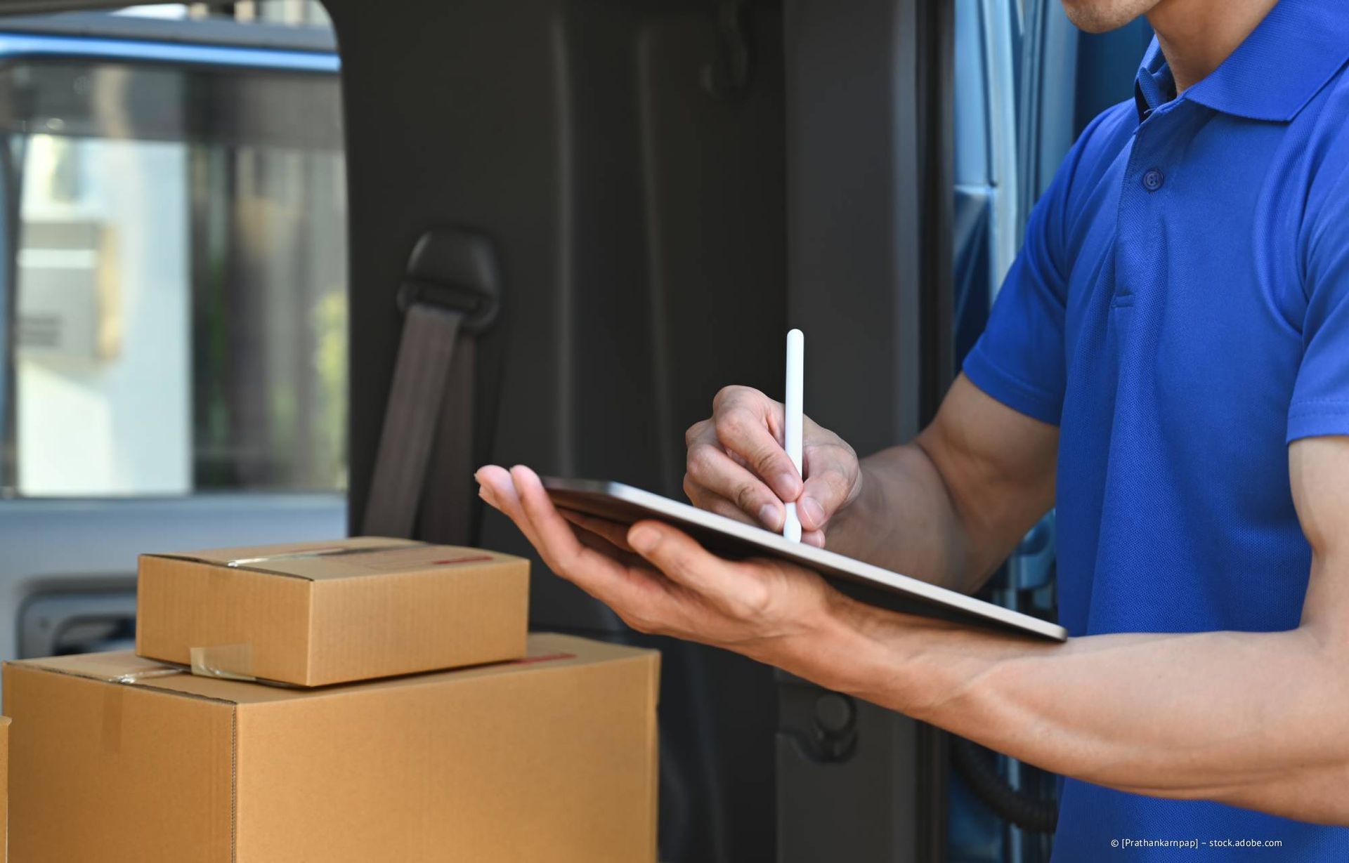 Ein Lieferant in blauer Uniform benutzt ein Tablet in der Nähe von Kartons in einem Lieferwagen.