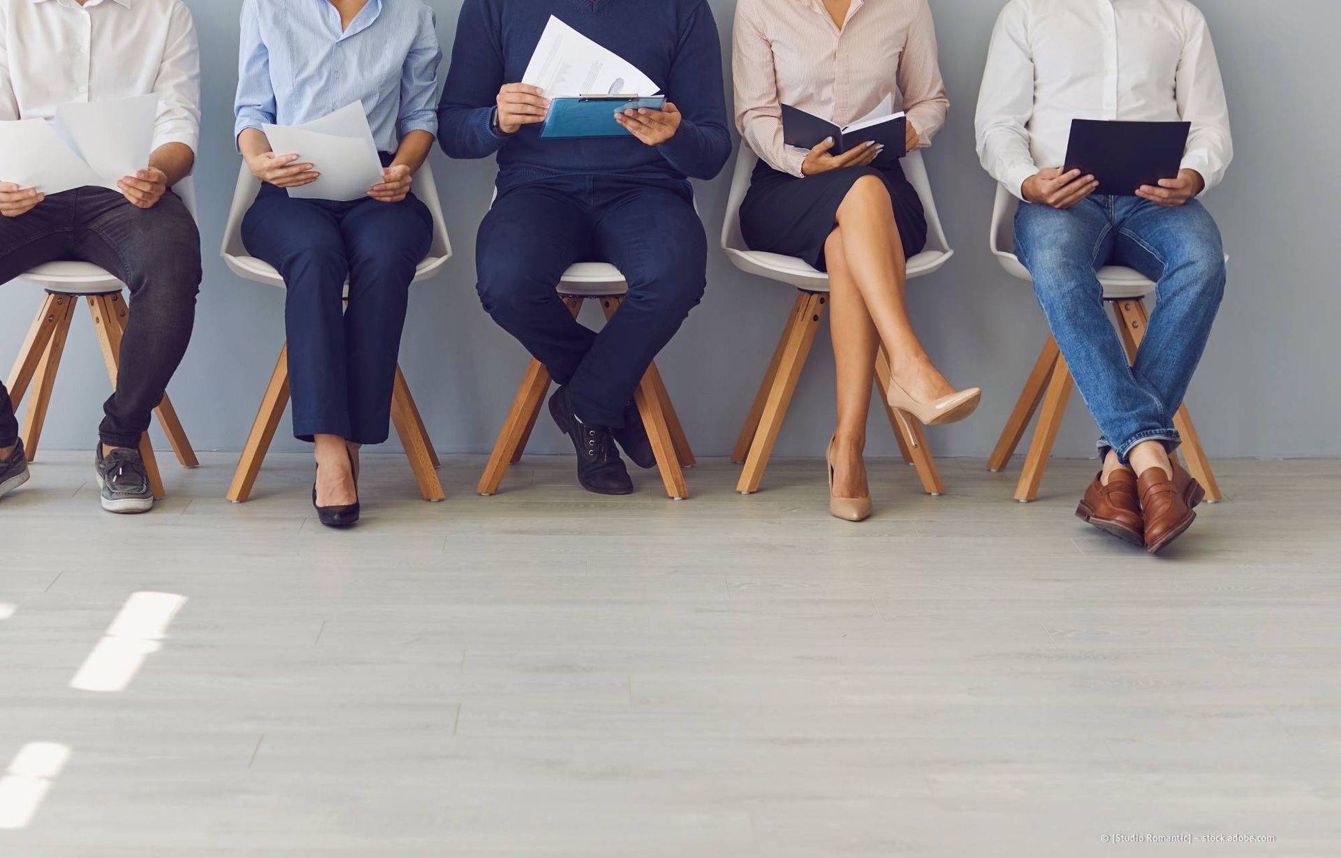 Fünf Personen sitzen auf Stühlen, halten Papiere oder Tablets in den Händen und warten in einem hell erleuchteten Raum.