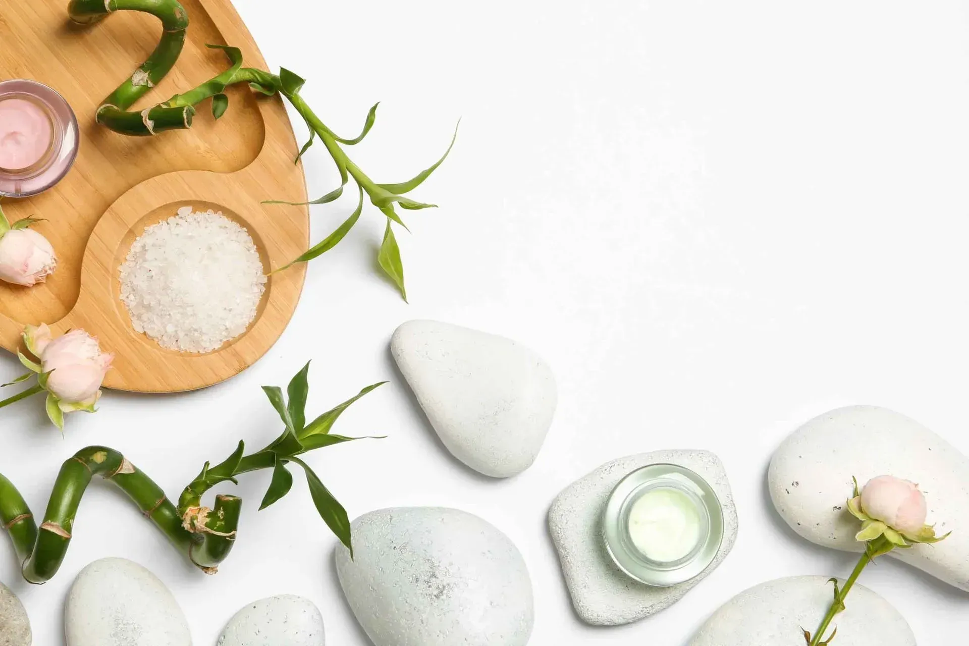 Bodegón de spa con piedras blancas, hojas verdes y cuencos de madera con sales de baño sobre un fondo claro.
