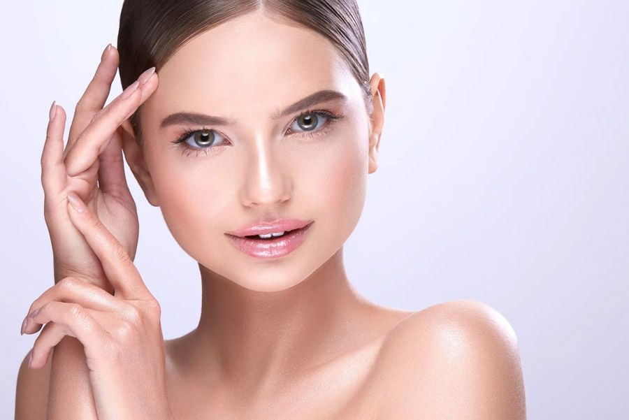 Mujer de piel suave, manos cerca de la cara, mirando a la cámara, fondo claro.