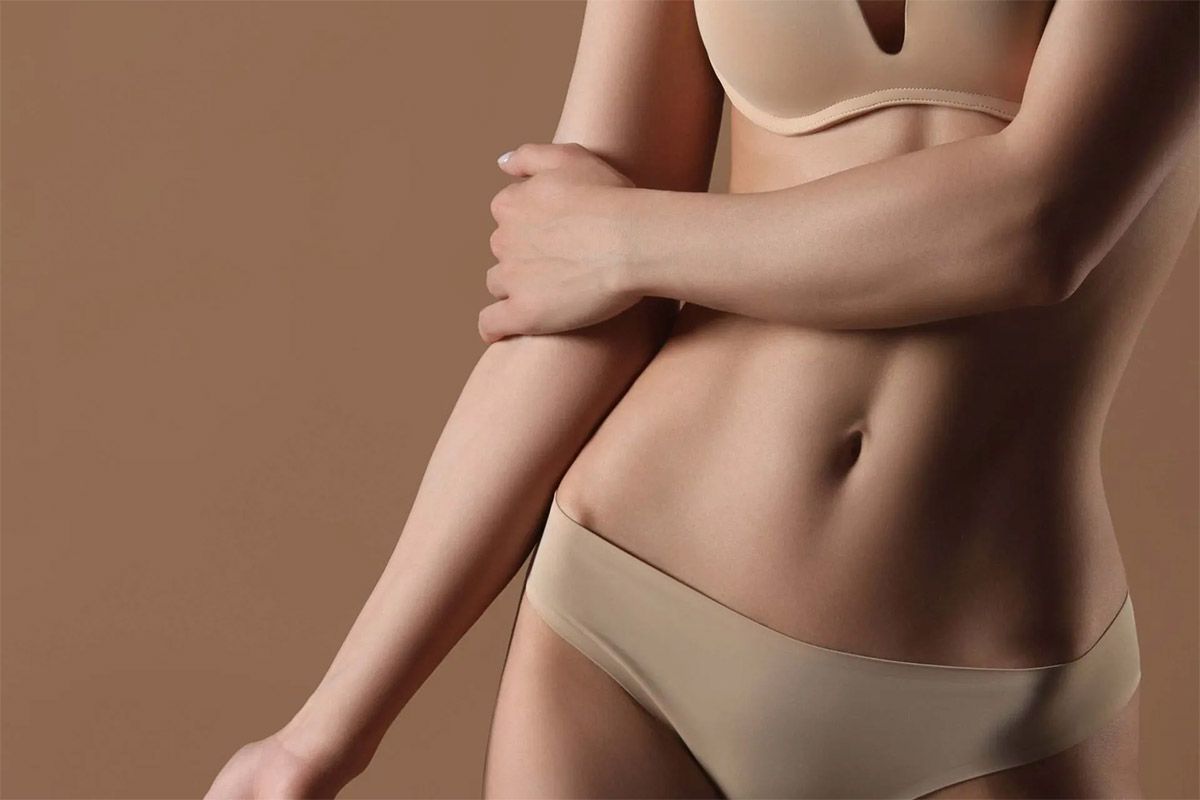 Mujer en lencería beige, con los brazos cruzados y el torso expuesto sobre un fondo color canela.