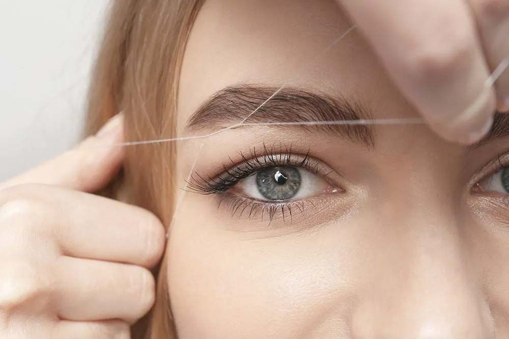 Depilación de cejas con hilo: el ojo de una persona se enmarca mediante una ceja que se delinea con un hilo sostenido por dos manos.