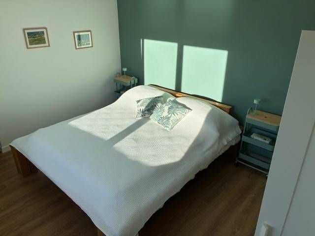 Schlafzimmer mit Doppelbett, türkisfarbener Akzentwand und Holzbodenoptik, mit zwei kleinen Tischen und gerahmten Kunstwerken.